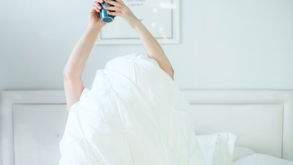 A person holding a coffee mug while relaxing in bed, covered with a white sheet.